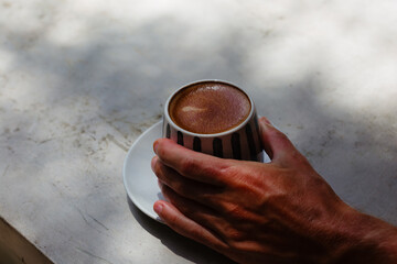 A man's hand holds delicious coffee in a ceramic mug on a concrete background. Cappuccino with brown foam against the background of sunlight. - obrazy, fototapety, plakaty