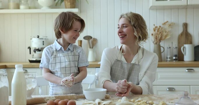 Cheerful cute little son boy helping mom to shape dough in kitchen, sprinkling with white flour powder. Happy mother and kid baking dessert, pastry together, talking, laughing, cooking at floury table