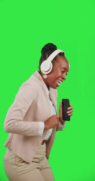 Black woman, dance with mobile phone and headphones on green screen, listen to music with fun and rhythm. Professional female with online streaming, tech and radio with freedom on studio background
