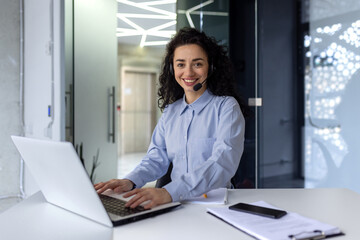 Portrait of successful hispanic woman at workplace inside office, woman with video call headset smiling and looking at camera, support worker working with laptop, help desk.