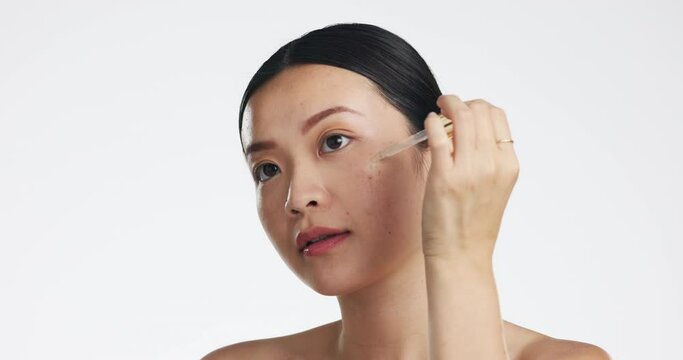 Asian woman, serum and retinol on face for beauty, skincare or cosmetics on white studio background. Happy female model applying oil dropper to skin for hydration, moisturizer or facial treatment
