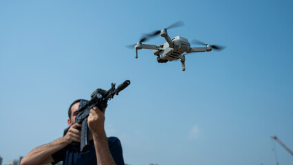 A man aims to shoot a rifle at a flying drone against a blue sky. 