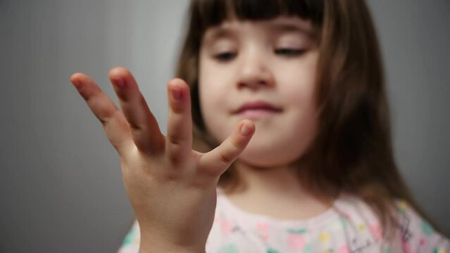 Isolated portrait of a little girl on a gray background who counts to five on her fingers right hand. A cute child smiling and counts from 1 to 5 fingers looking at his hand.