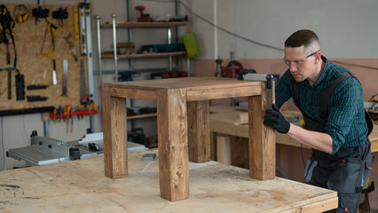 Male carpenter finishing work on wooden table in workshop. 