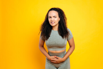 Unhealthy brazilian or hispanic young woman in sportswear, keeps hands on a stomach, demonstrating...
