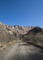 A dusty road in the autumn mountains on a sunny November morning goes into the distance, against the backdrop of rocky steep peaks and ridges