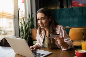 A young woman uses her credit card to shop for office supplies online on her laptop while sitting at a local café