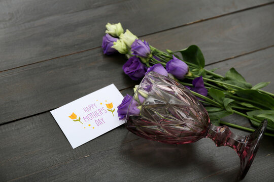 Greeting card with text HAPPY MOTHER'S DAY, glass and eustoma flowers on black wooden background, closeup