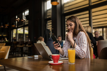 Woman using phone while working remotely on her laptop in a crowded coffee shop