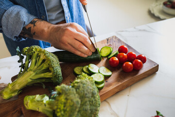 A chef's hands chop and slice fresh vegetables (cucumber, cherry tomato, broccoli) on a wooden board