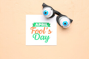 Paper with text APRIL FOOL'S DAY and funny eyeglasses on beige background