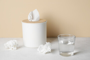 Fototapeta na wymiar Glass of water with tissue box on table near beige wall. Allergy concept