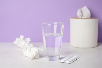 Fototapeta na wymiar Glass of water with pills and tissue box on table near lilac wall. Allergy concept