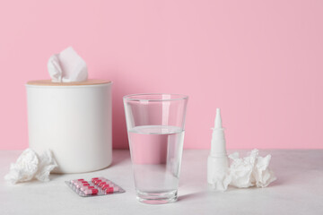 Fototapeta na wymiar Glass of water with pills, nasal drops and tissue box on table near pink wall. Allergy concept