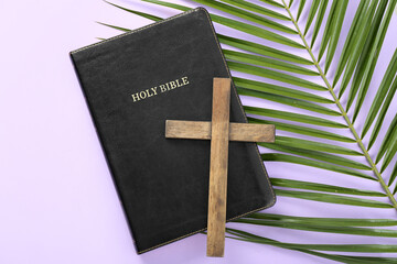 Wooden cross with Holy Bible and palm leaf on lilac background. Good Friday concept