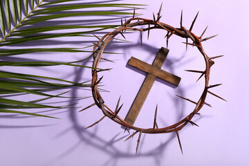 Fototapeta na wymiar Wooden cross with crown of thorns and palm leaf on lilac background. Good Friday concept