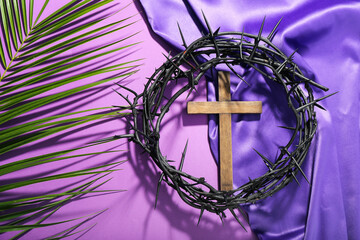 Wooden cross with crown of thorns, palm leaf and purple fabric on violet background. Good Friday...