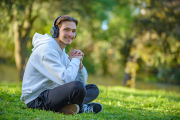 Naklejka na ściany i meble Portrait of young man in headphones listen to the music outdoors. Young smiling guy with headphones sitting on green lawn in the park and relaxing. Happy young man rests listening the music on nature