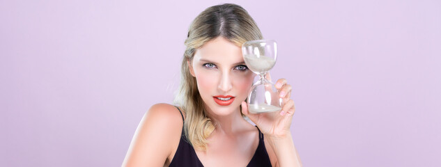 Alluring beauty model holding hourglass in forever young beauty concept of anti-aging skincare treatment for woman. Beautiful caucasian women portrait with perfect makeup in pink isolated background. - obrazy, fototapety, plakaty