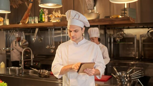 Portrait of beautiful successful male chef preparing for cooking using tablet online scrolling. Handsome famous cook wearing white uniform at fancy restaurant kitchen. Cooking concept.