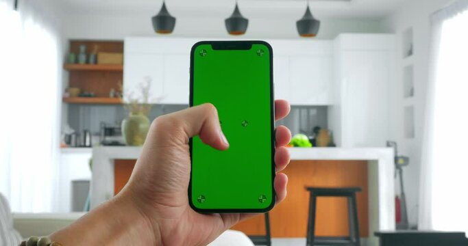 Close up man's hand holding mobile phone with green screen indoors on modern kitchen background, doing swipe up gesture on chroma key of modern mobile phone on home kitchen background