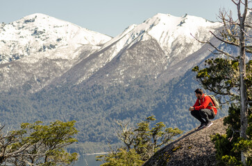 hombre en la cima de un cerro, sobre una roca, observando el paisaje de lagos y montañas nevadas