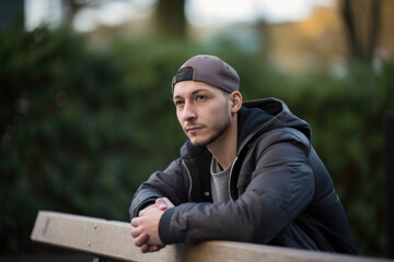 Obraz premium Thoughtful portrait of a young man with a baseball cap and a bomber jacket, sitting on a bench in a park, generative ai