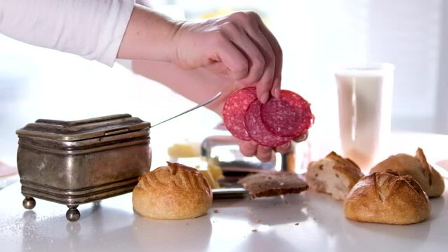 preparing sandwich close-up female hands put Three pieces of sausage on a bun with butter stands a glass of milk large silver sugar bowl delicious pastries on a white table crumbs