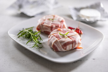 Camembert or brie cheeses wrapped in bacon with fresh rosemary and chilli on a white plate