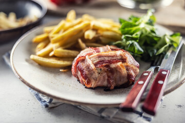 Bacon wrapped meat served with french fries and salad on a plate with cutlery