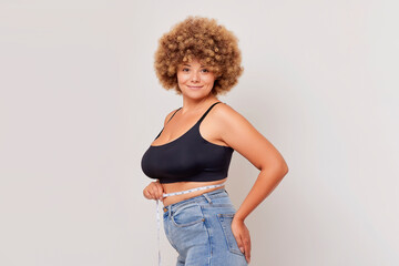 Cute young curly woman in black top with measurer on her waist stands half turned on light...