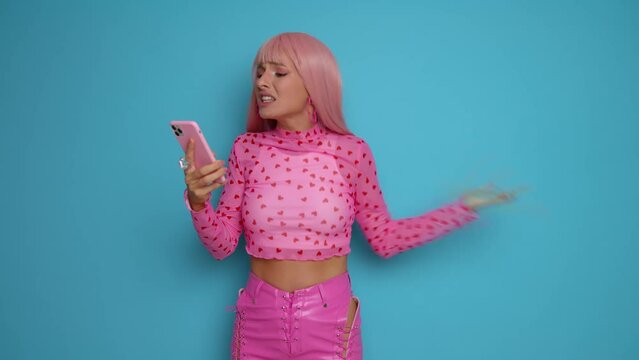 Young woman in pink clothes standing on blue studio background, reads something unpleasant on the phone and waves her hand indignantly showing disappointment, copy space. 
