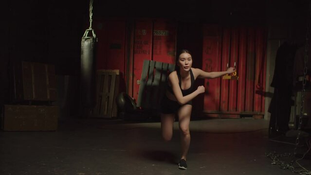 Determined active attractive sporty fitness Asian woman practicing cardio workout, for balance, agility and mobility, performing skater jump exercise, building lower body strength in dark garage gym.