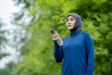 Asian cute muslim woman wearing sportswear and a blue hijab doing exercise and being tired and drinking water outdoors at the park in the morning with a fresh feeling.