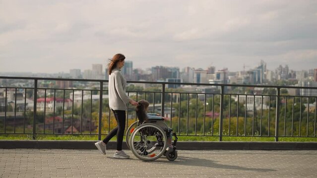 Mother shows city panorama to little daughter sitting in wheelchair. Cheerful woman and girl with paralysis enjoy walk in park on sunny spring day
