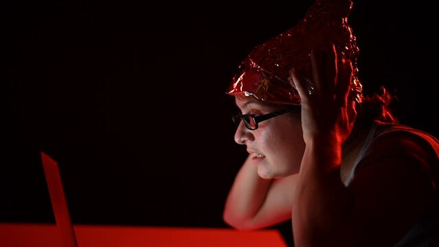 Paranoid caucasian woman typing on a computer wearing a tinfoil cap. Conspiracy theory.