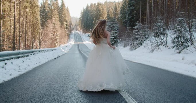 Beautiful Caucasian bride in wedding dress long hair walking running dancing on asphalt road among winter snowy pine forest. Gorgeous young blonde woman alone in dress in winter in Ukraine slow motion