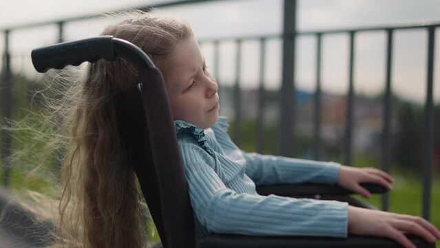 Preschooler girl with paralysis sits in wheelchair looking at city on blurred background. Tranquil girl with disability enjoys sunny spring weather closeup