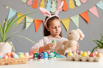 Image of adorable cute little girl dressed in rabbit ears sitting at table, holding her soft toy and showing her beautiful green Easter eggs to her fluffy friend, posing against decorated gray wall