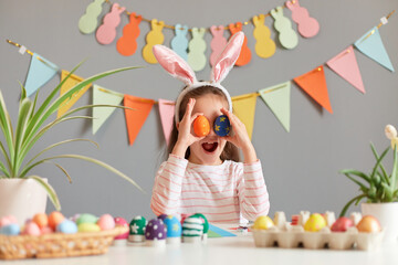 Portrait of amazed surprised little girl dressed in rabbit ears sitting at table, covering her eyes...