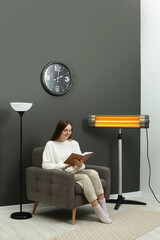 Young woman reading book on armchair near electric infrared heater at home