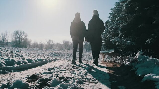 Back view of two women walking hand in hand along a snowy path near the winter forest, slow motion. Girlfriends walking by snow against the bright sun. Concept of a loving couple, lesbian, LGBT