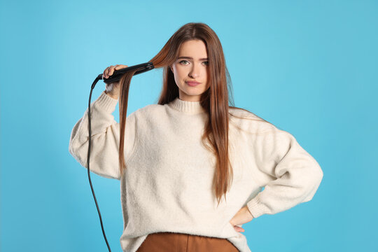 Upset young woman with flattening iron on light blue background. Hair damage