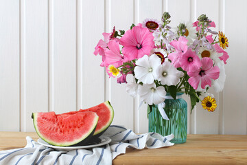 bouquet of garden flowers in a glass vase and a cut watermelon on the table