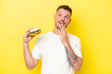 Young caucasian man holding a burger isolated on yellow background looking up while smiling