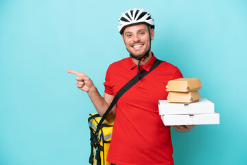 Young caucasian man with thermal backpack and holding fast food isolated on blue background pointing finger to the side
