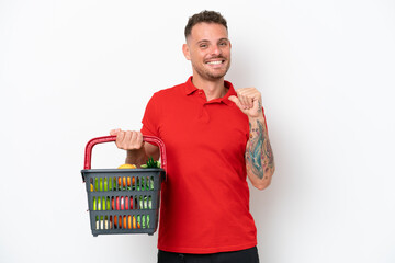 Young caucasian man holding a shopping basket full of food isolated on white background proud and self-satisfied