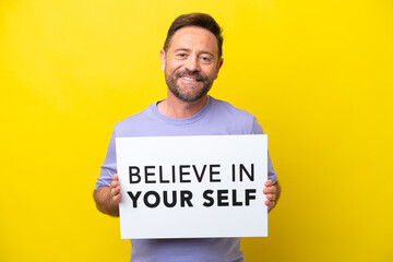 Middle age caucasian man isolated on yellow background holding a placard with text Believe In Your Self with happy expression