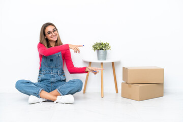 Young caucasian woman sitting on the floor among boxes pointing finger to the side and presenting a product