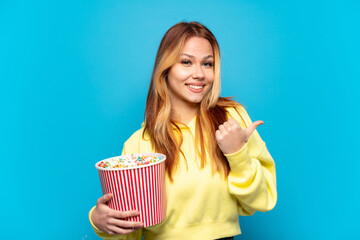 Teenager girl holding popcorns over isolated blue background pointing to the side to present a...